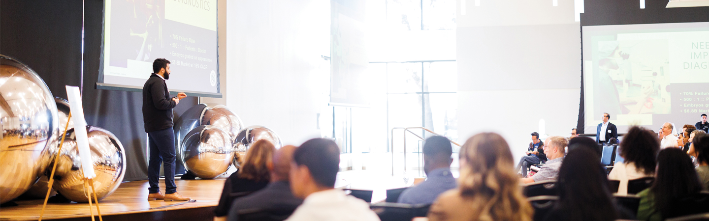 person standing on stage speaking to a large group of people at a pitch event